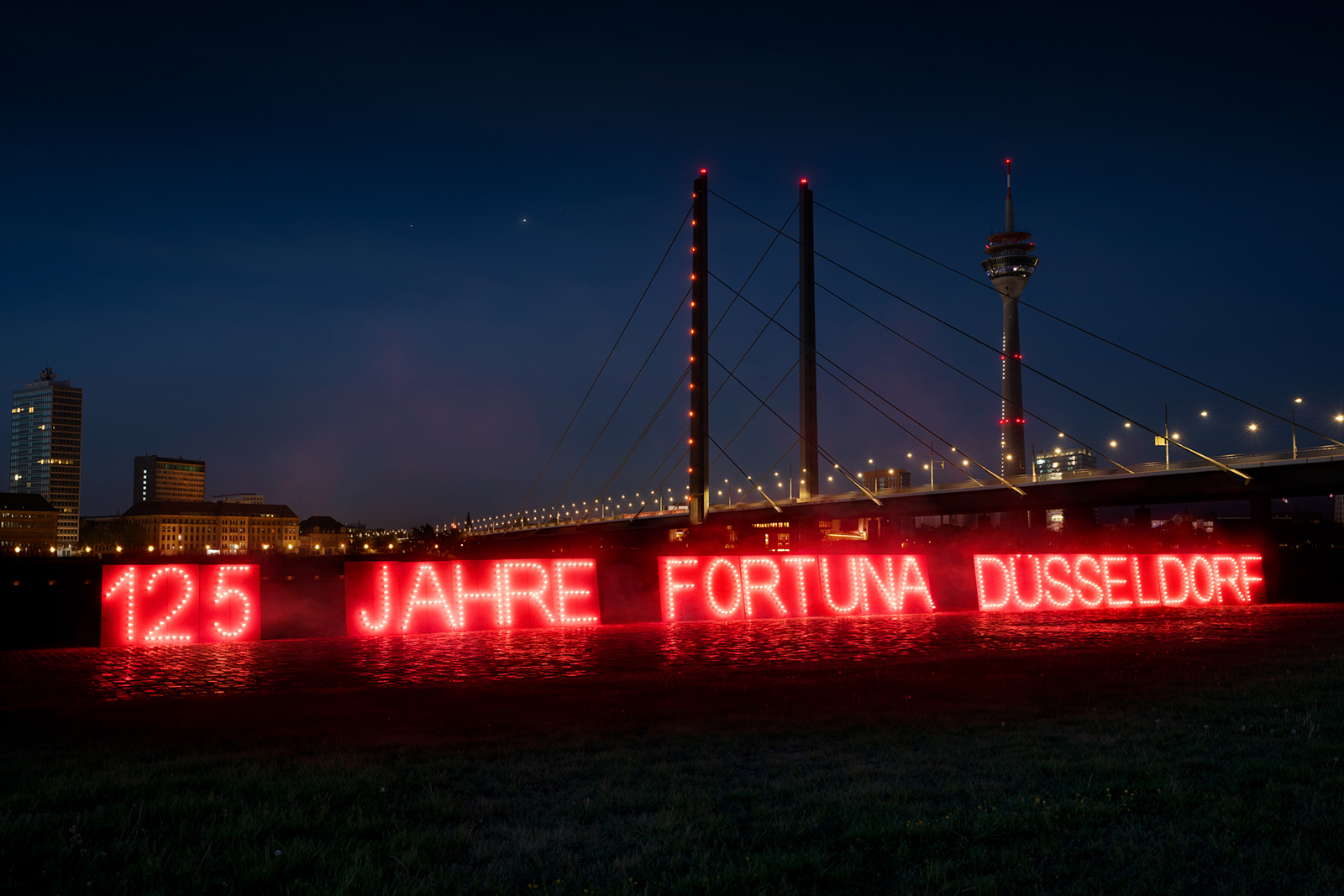 rot leuchtende Schrift: "125 Jarhe Fortuna Düsseldrf" vor der Rheinbrücke bei Nacht
