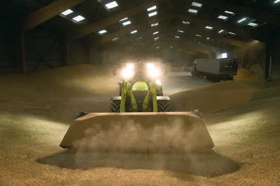 Traktor mit großer Schaufel voll Korn in einer Lagerhalle voll Korn, Lkw im Hintergrund