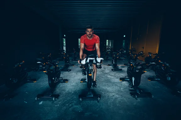 Männliche Person in Sportkleidung auf einem Fahrrad in einem Raum voller Fahrräder, sehr dunkel