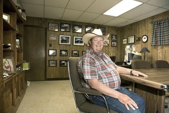 Mann mit Cowboyhut sitzt in einem Raum an einem Tisch, Bilder im Hintergrund an der Wand