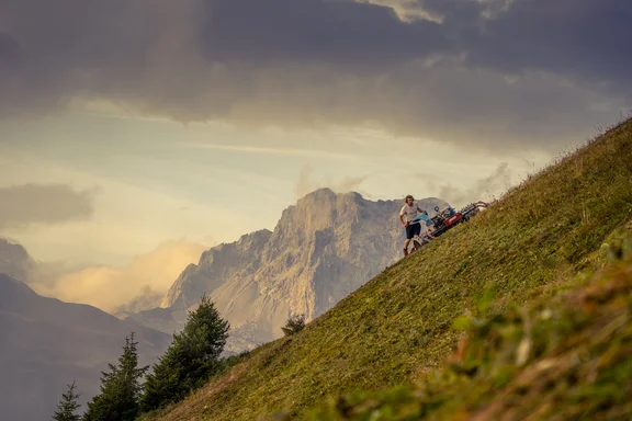 Person mit Grasschneidemaschine auf einer Wiese an einem Berg, Berge im Hintergrund