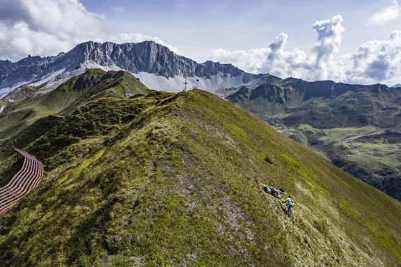 Mann, der auf einem Berg ein Gerät über die Wiese schiebt, Hintergrund mehr Berge