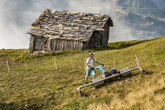 Eine Holzhütte und ein Mann, der davor auf einer Wiese am Berg Gras auflockert mit einem großen Gerät