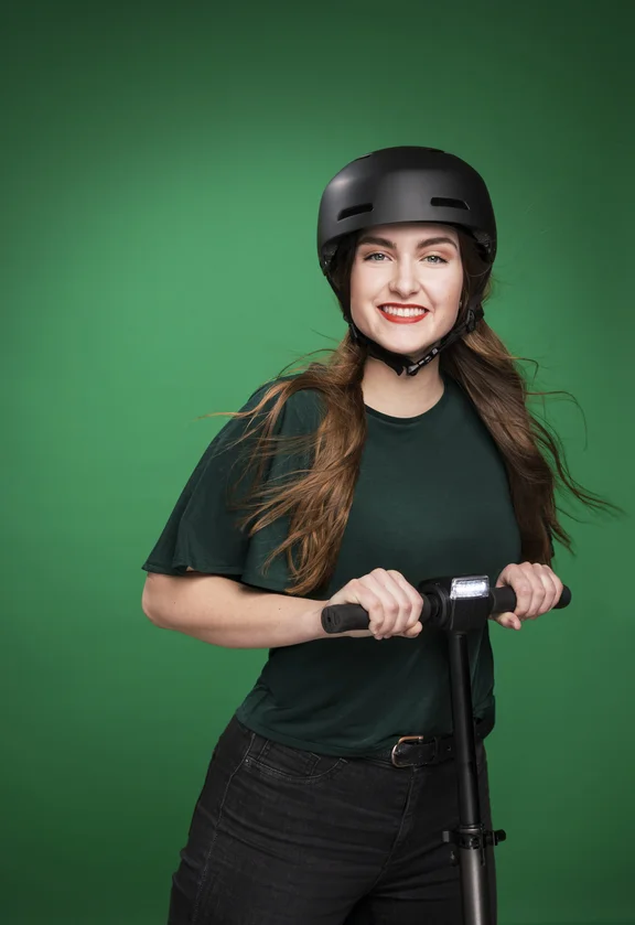 Weibliche Person mit dunkelgrünem T-Shirt vor grüner Wand auf einem Cityroller, mit Helm