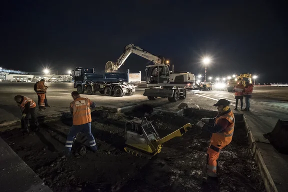 Bauarbeiter, die an einem aufgerissenen Parkplatz arbeiten, Bagger im Hintergrund, bei Nacht