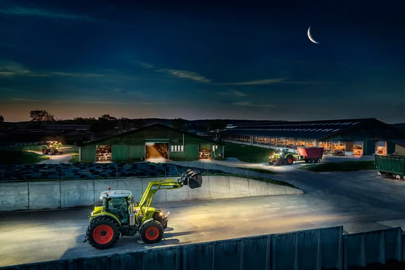 drei Traktoren, die neben zwei Kuhställen fahren und Maschinen ziehen, bei Nacht