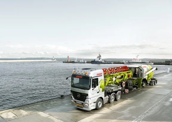 Lkw beladen mit einem Mähdrescher und einem Mähwerk steht am Hafen, links Wasser, rechts Lkw