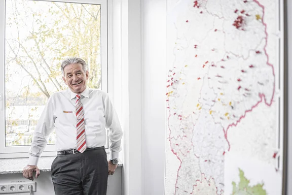 Älterer Mann in Hemd steht am Fenster, Landkarte an der Wand daneben