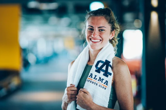 Portrait Frau in Sportkleidung und Handtuch um dem Hals, Hintergrund ist ein unscharfes Sportstudio