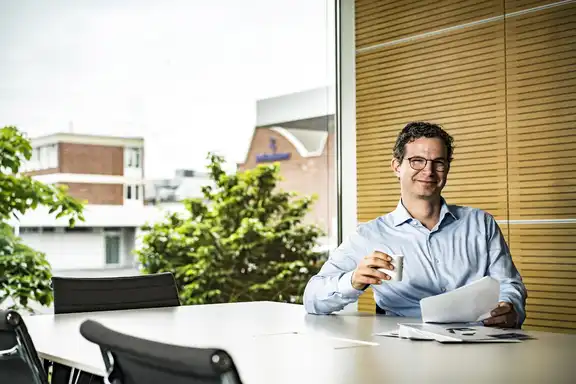 Mann mit Zetteln und Kaffee an einem Tisch, rechte Wand aus Holz, hinter dem Mann ein großes Fenster