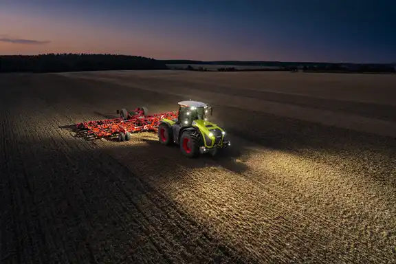 Trecker zieht großen Grubber über ein Feld, Blick von schräg oben, in der Dämmerung