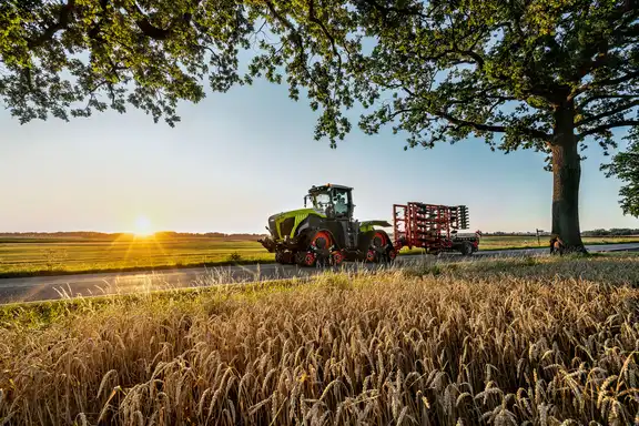 Landmaschine "Raupe" zieht Grubber über eine Straße, Felder auf beiden Seiten, Bäume am Straßenrand