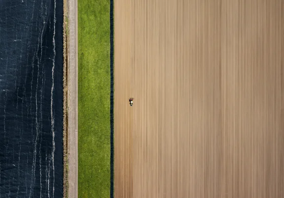 Ggroßes Feld mit kleinem Trecker rechts, Grassstreifen in der Mitte, Wasser links