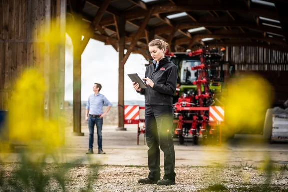 Frau in Arbeitskleidung mit Tablet in der Hand vor einem Trecker