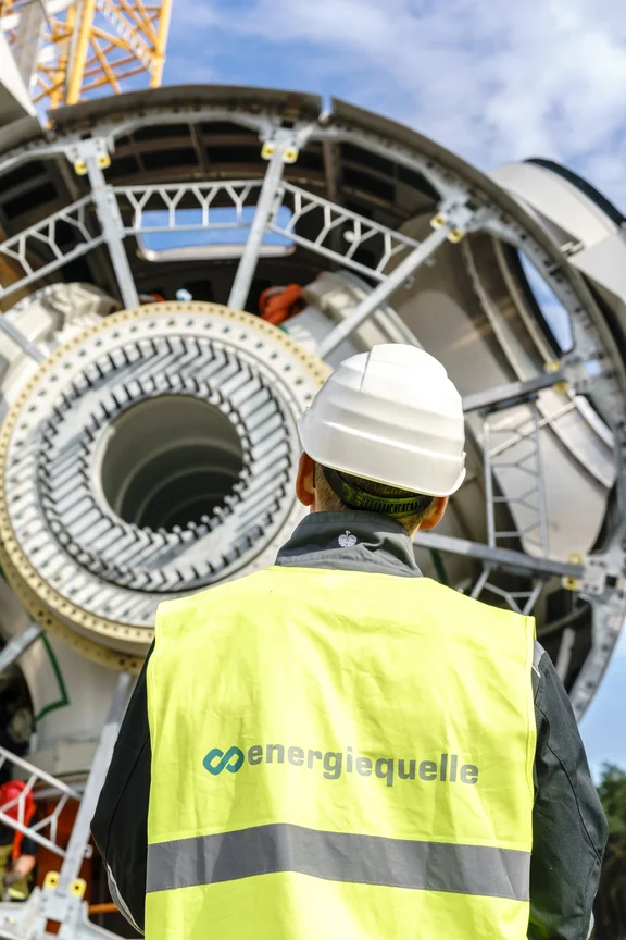 Mann in Warnweste mit Helm von hinten steht vor einem Windradteil