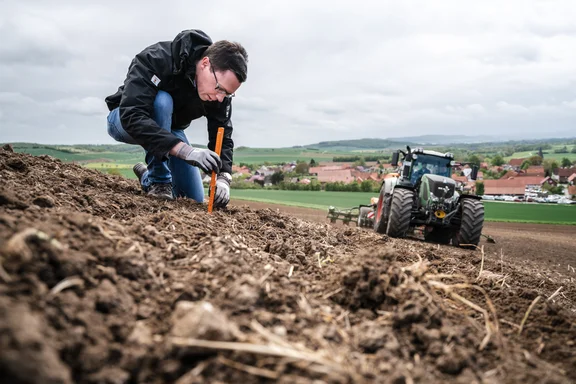 Mann, der auf einem Acker einen Stab ins Feld steckt, rechts daneben ein Traktor, Hintergrund: Felder, ein paar Häuser