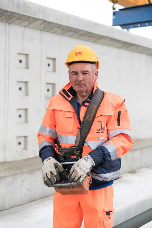 Arbeiter in orangener Kleidung mit Helm, Steuergerät in der Hand, vor einer Betonmauer