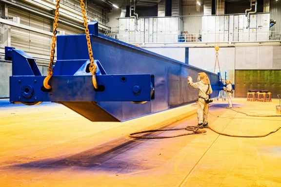 Zwei Personen in Schutzkleidung lackieren in einer Halle ein großes Teil in blau