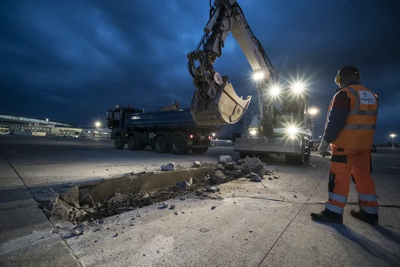 Bagger reißt Parkplatz auf, Lkw im Hintergrund, Person in Arbeitskleidung steht daneben