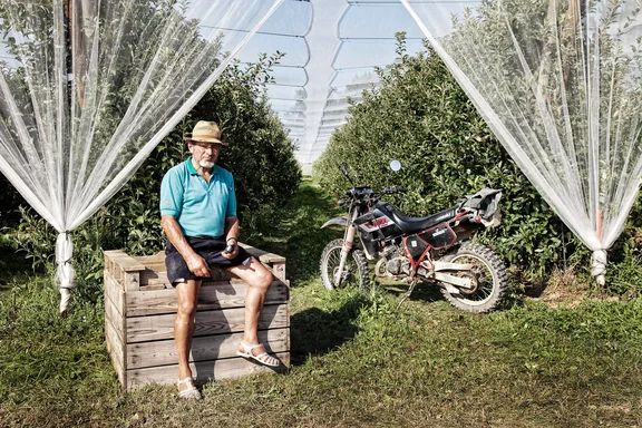 Apfelbauer sitzt auf einer Holzkiste in mitten eines Apfelfeldes, neben ihm ein Motorrad