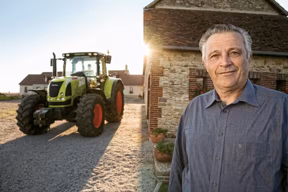 Mann rechts vor einem Haus, links steht ein grüner Trecker