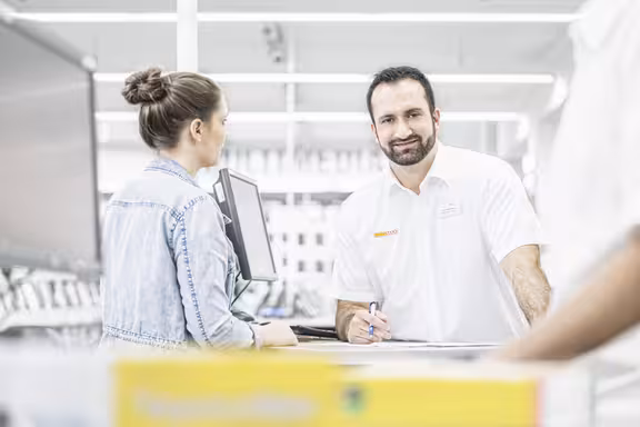 Mann steht an einer Kasse und redet mit einer Kundin