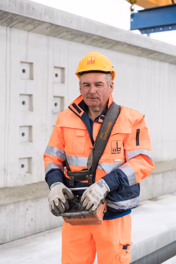 Arbeiter in orangener Kleidung mit Helm, Steuergerät in der Hand, vor einer Betonmauer