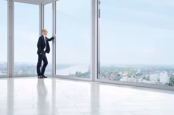 Mann, der in einem Raum aus Fenstern an einem Fenster lehnt und auf die Stadt herunter schaut