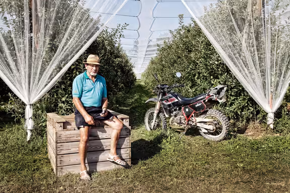 Apfelbauer sitzt auf einer Holzkiste in mitten eines Apfelfeldes, neben ihm ein Motorrad