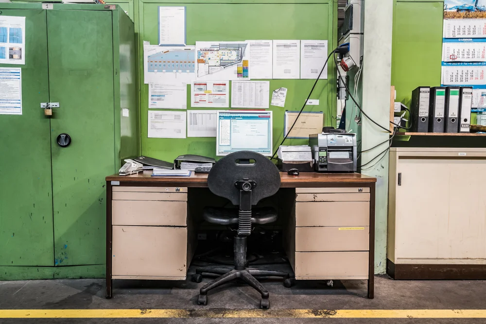 Schreibtisch mit Schubladen auf beiden Seiten mit Bürostuhl vor einer Grünen Wand, grüner Metallschrank links, Kommode mit Ordnern rechts